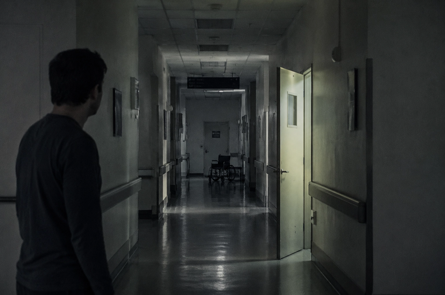Uomo fermo in un corridoio d’ospedale poco illuminato, con una porta socchiusa e una sedia a rotelle vuota in fondo