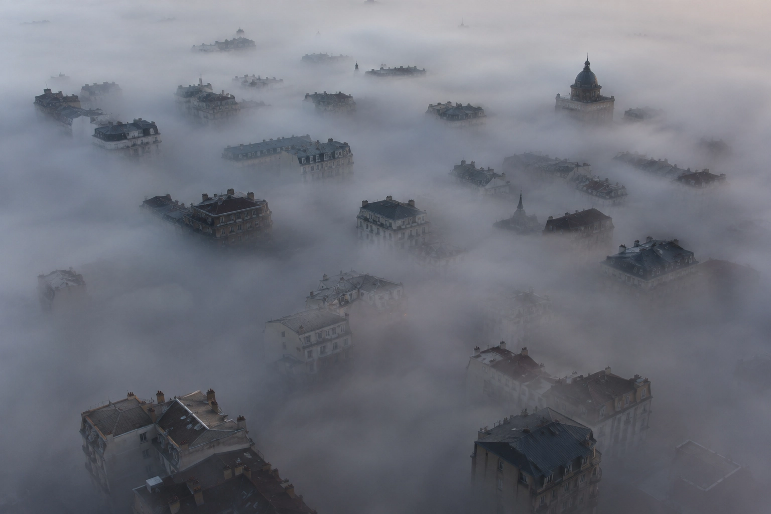 Veduta aerea di un quartiere urbano coperto da una nebbia molto fitta, con tetti, campanili e piani superiori degli edifici che emergono sopra lo strato bianco